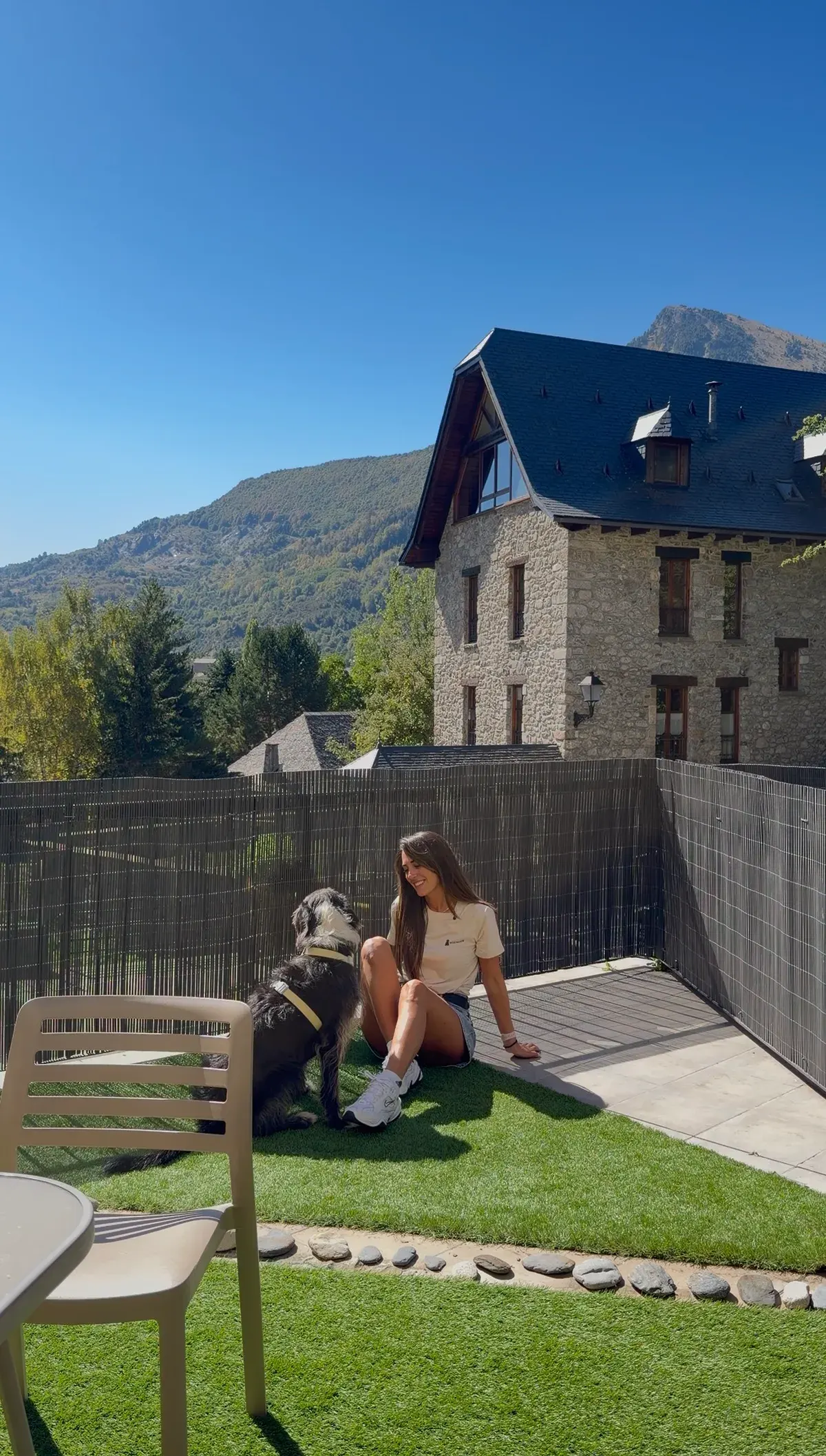 Kelly sentada con su perro en el jardín vallado de un alojamiento de montaña, junto a una casa de piedra